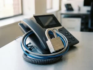 Stahlvorhängeschloss auf einem IP-Tischtelefon mit umschlungenem Ethernet-Kabel, symbolisiert VoIP-Sicherheit im Büro.